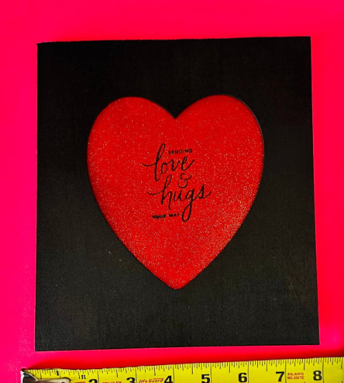 Red heart-shaped card with 'love & hugs' text on a black background, measured against a pink border.