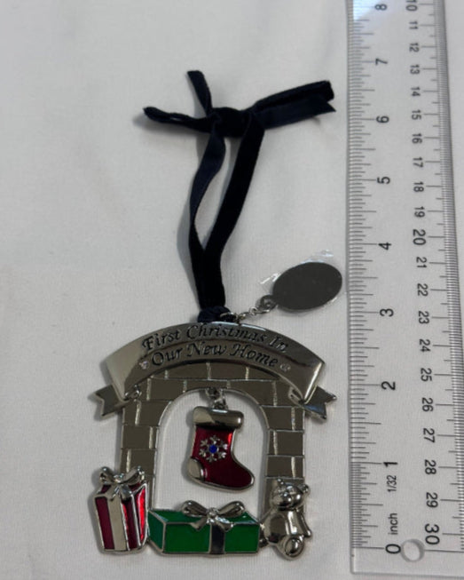 Decorative Christmas ornament with a stocking, presents, and a baby figure, hanging on a black ribbon next to a ruler for scale.