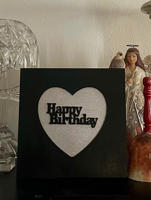 Black sign with a white heart and 'Happy Birthday' text, placed on a surface with decorative items.