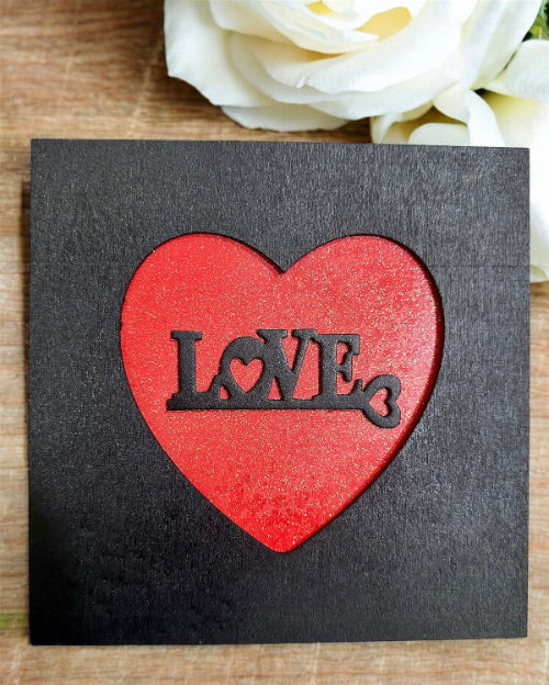 Black card with a red heart-shaped cutout featuring the word 'LOVE', placed on a wooden surface with white flowers.
