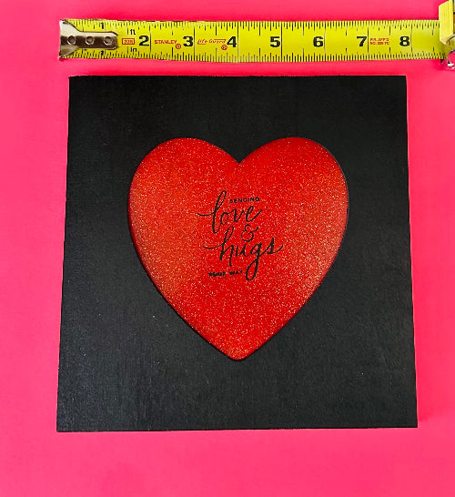 Red heart-shaped card with 'Sending love & hugs' text on a black background, measured against a pink border.