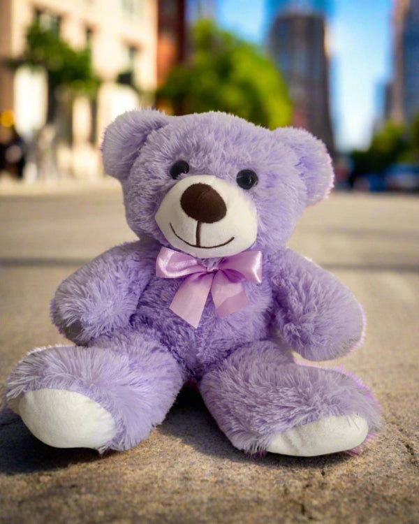 Purple teddy bear with a pink bow sitting on a concrete surface with a cityscape in the background.