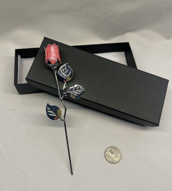 Decorative hair stick with pink rose and leaves on a gray surface with a black box.