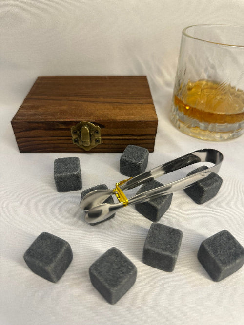 Whiskey stones, tongs, and a wooden box on a white surface with a glass of whiskey.