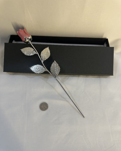 Silver rose with leaves on a white surface next to a black box.