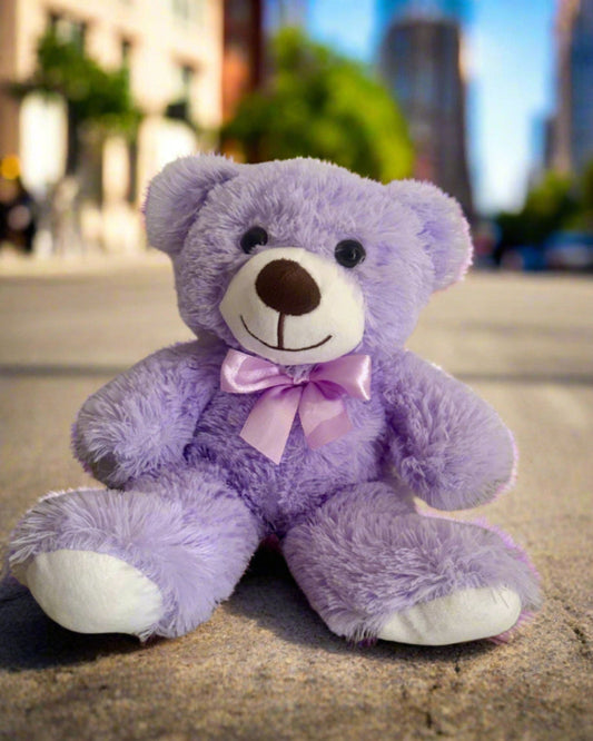 Purple teddy bear with a pink bow sitting on a concrete surface with a cityscape in the background.