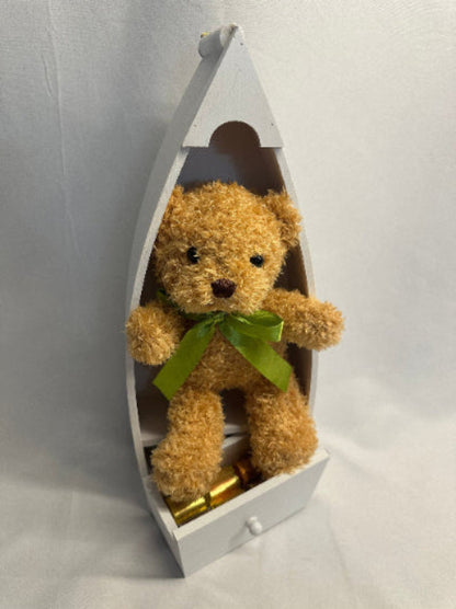Brown teddy bear with a green bow in a white boat-shaped box on a light gray background