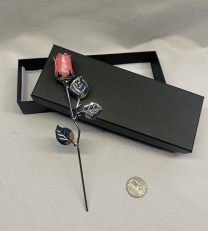 Decorative hair stick with pink rose and leaves on a gray surface with a black box.